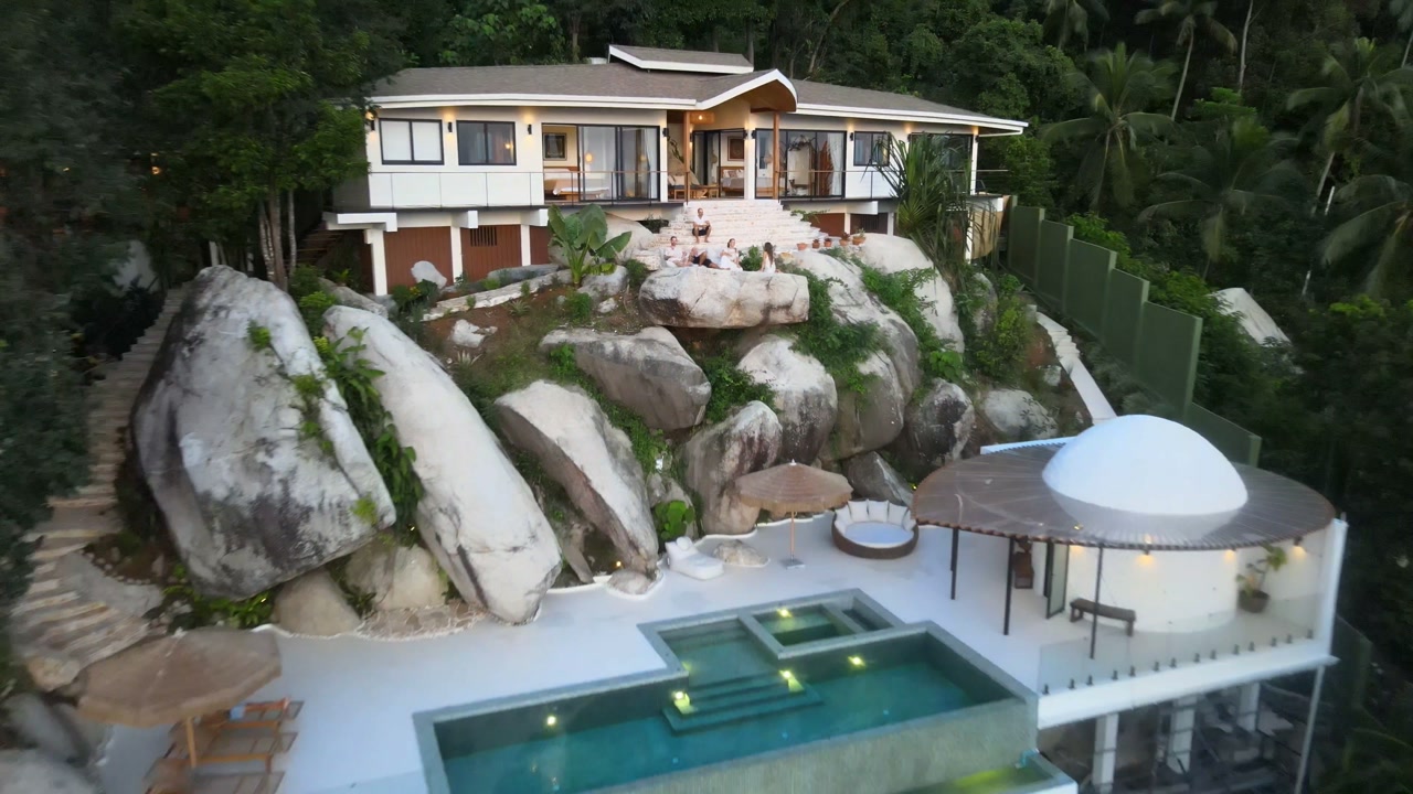 Villa Ullassa at dusk from the air: the four buildings lit up, guests on the stone steps, the pool and the domed spa glowing below