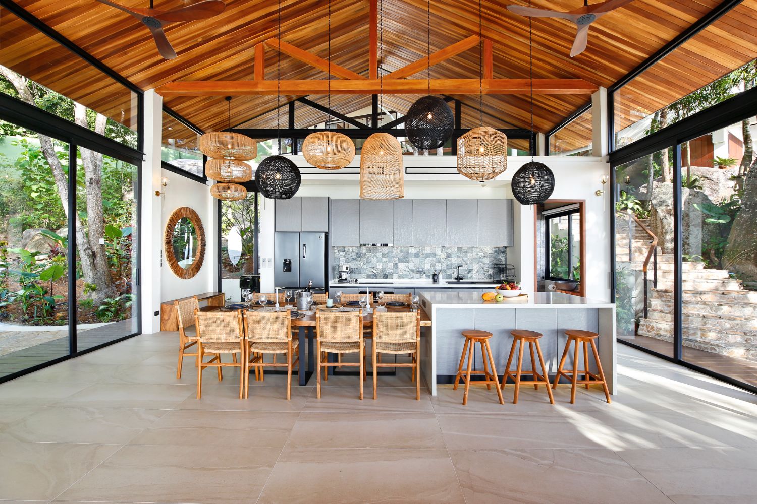 Open kitchen and long dining table under woven pendant lights, with glass walls on both sides of the pavilion