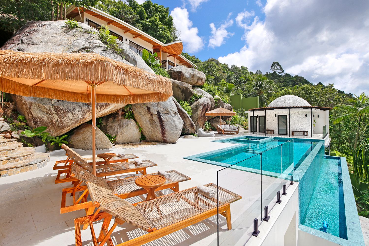 Teak sun loungers and thatched parasols on the pool terrace beside the boulders, jungle hills beyond