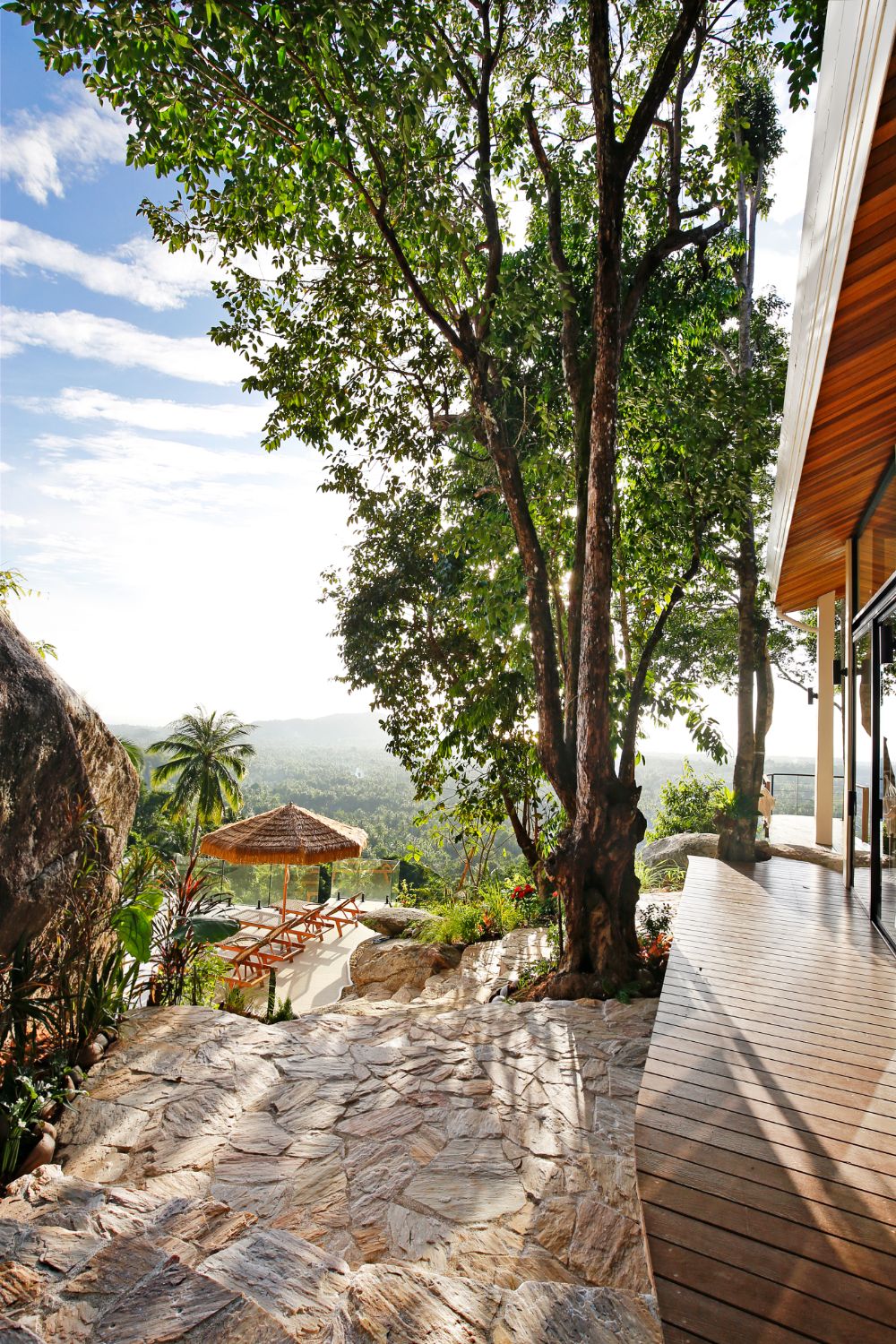 Stone-paved terrace under a large tree, the teak deck to one side and a parasol and loungers below
