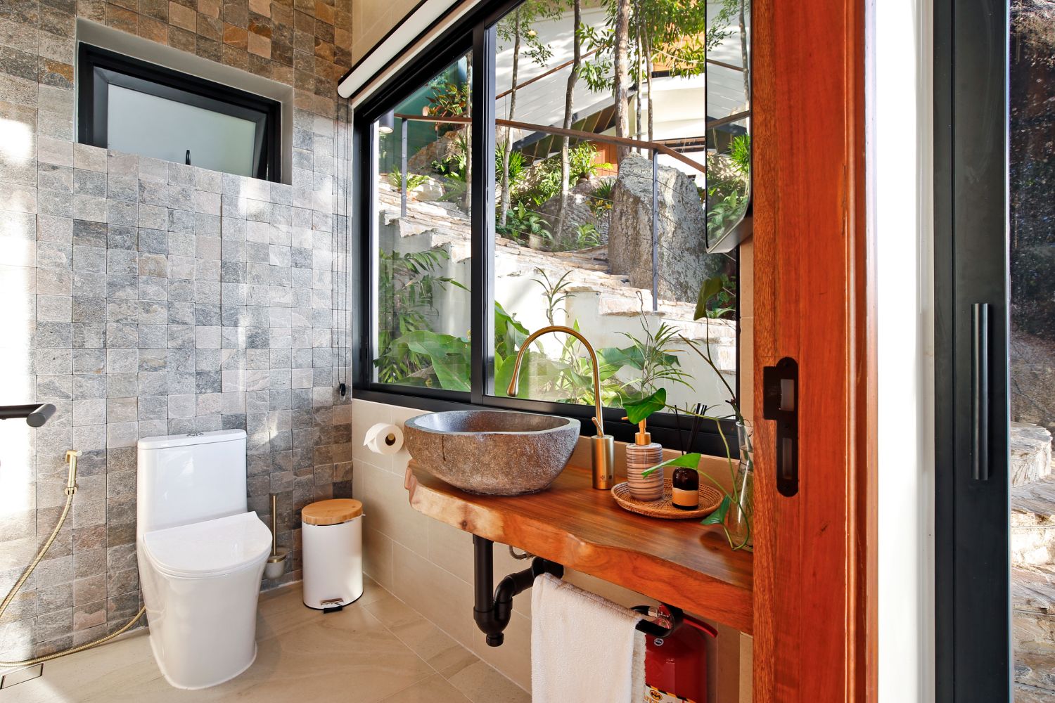 Guest bathroom with a stone basin on a teak counter and a window opening onto the planting outside