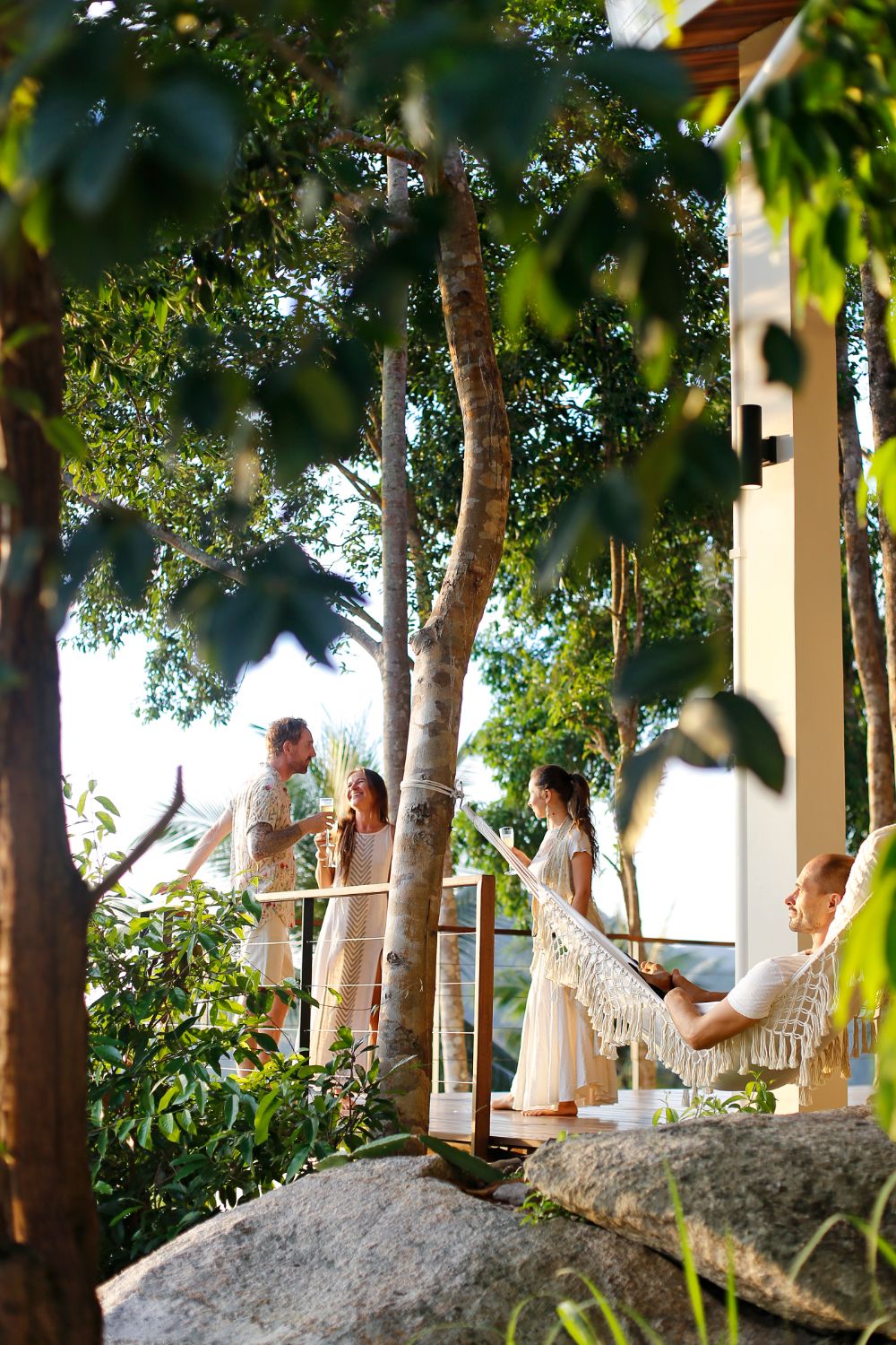 Guests at the terrace railing and in the hammock at golden hour, seen through the trees