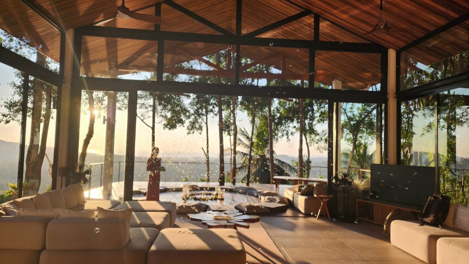The living pavilion at golden hour, low sun coming through the trees across the sofas and the long table