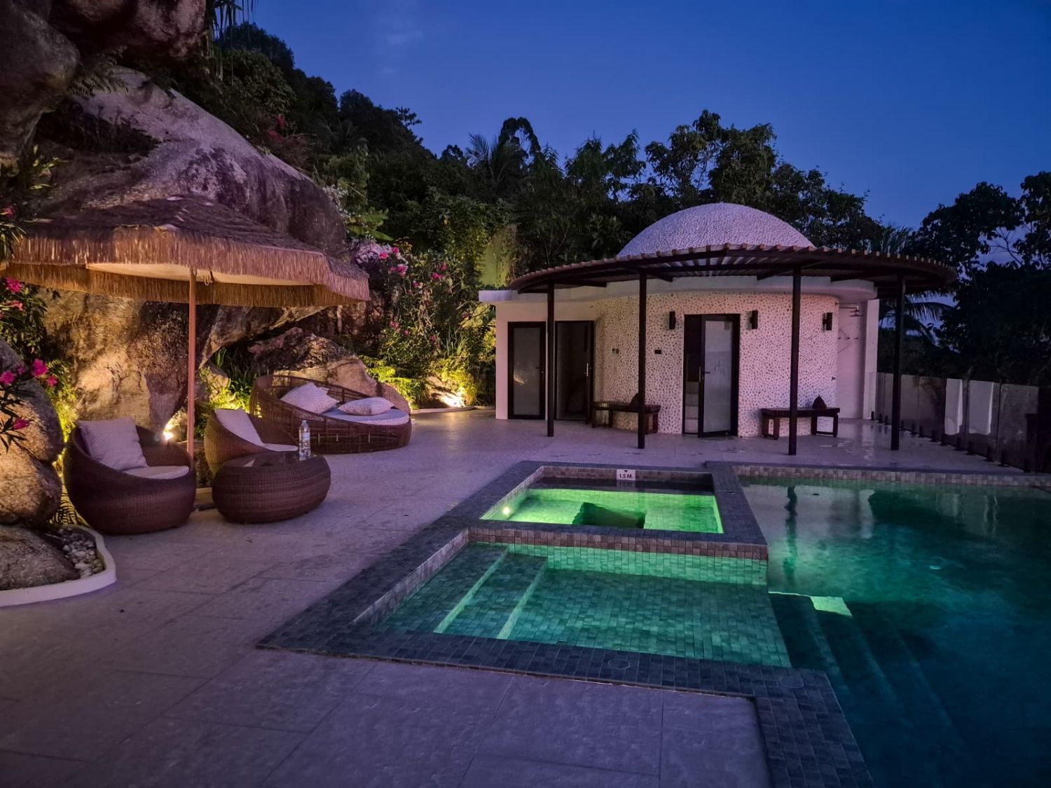 Pool and jacuzzi lit from within at night, beanbags and a thatched parasol on the terrace beside the dome