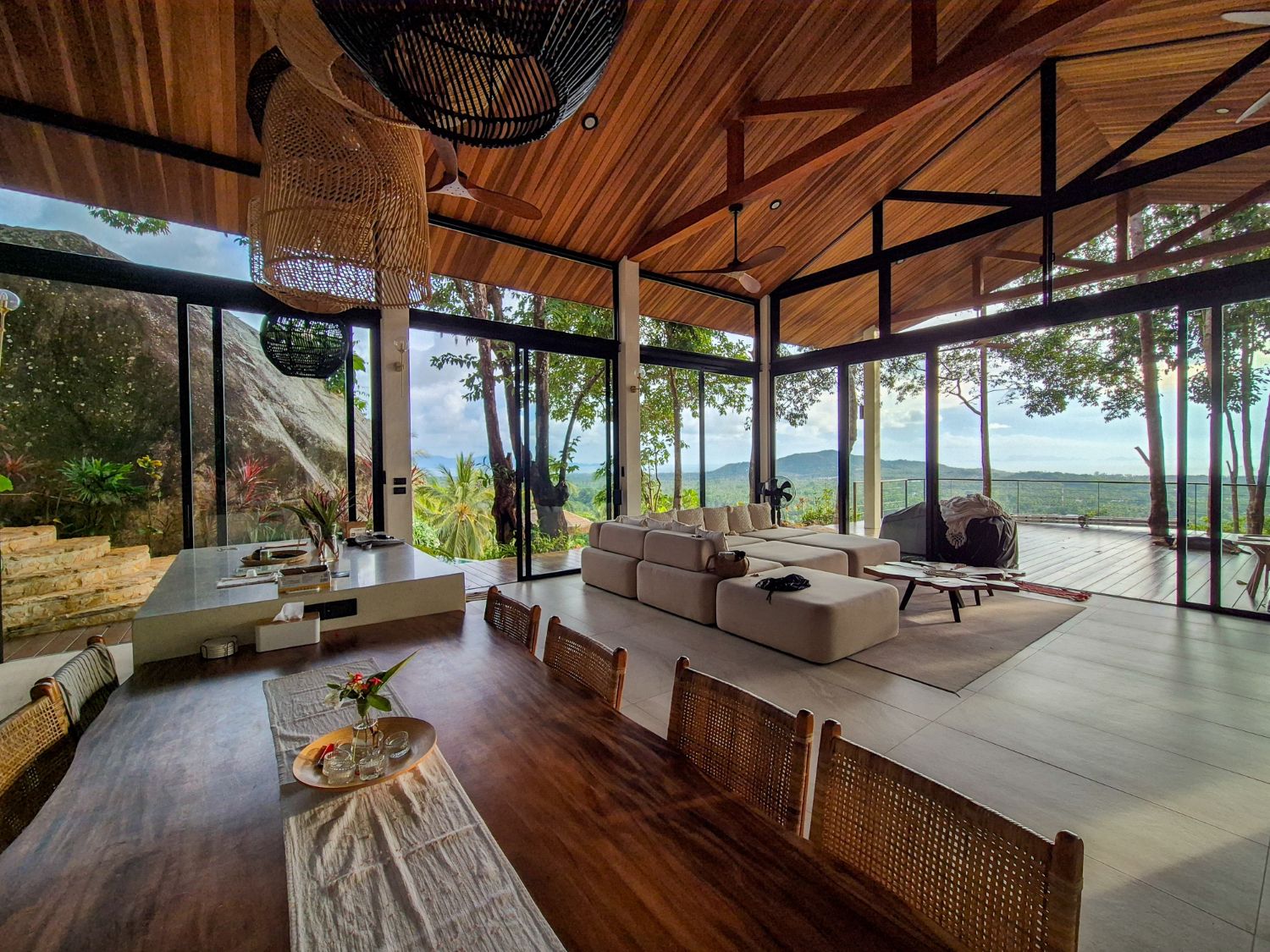 Living and dining pavilion with a dark woven pendant over the table, sofas beyond and the hills through the glass