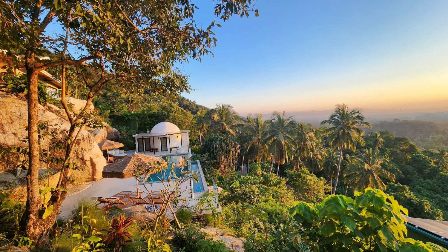 Golden-hour view from Villa Ullassa's hilltop over the pool, the domed building and the palms toward the horizon