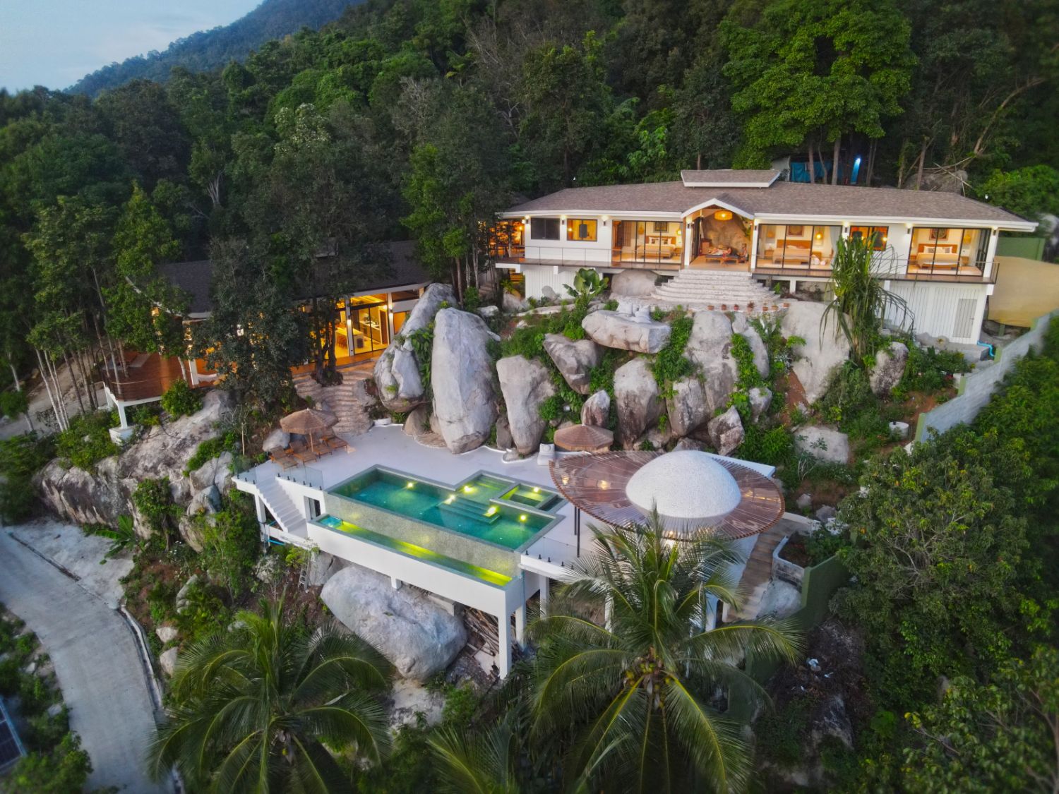 Aerial view at dusk of Villa Ullassa's four buildings, the pool and the domed spa set among granite boulders and palms on the jungle hillside