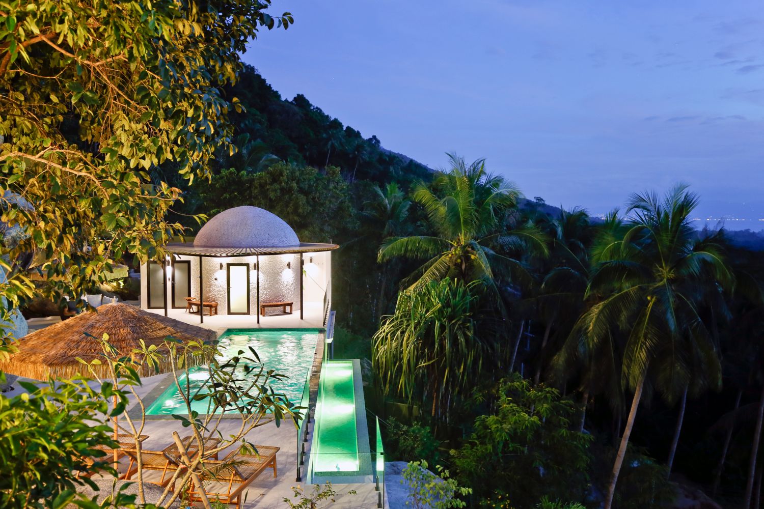 The domed building and the pool lit at dusk, palms rising around them on the hillside