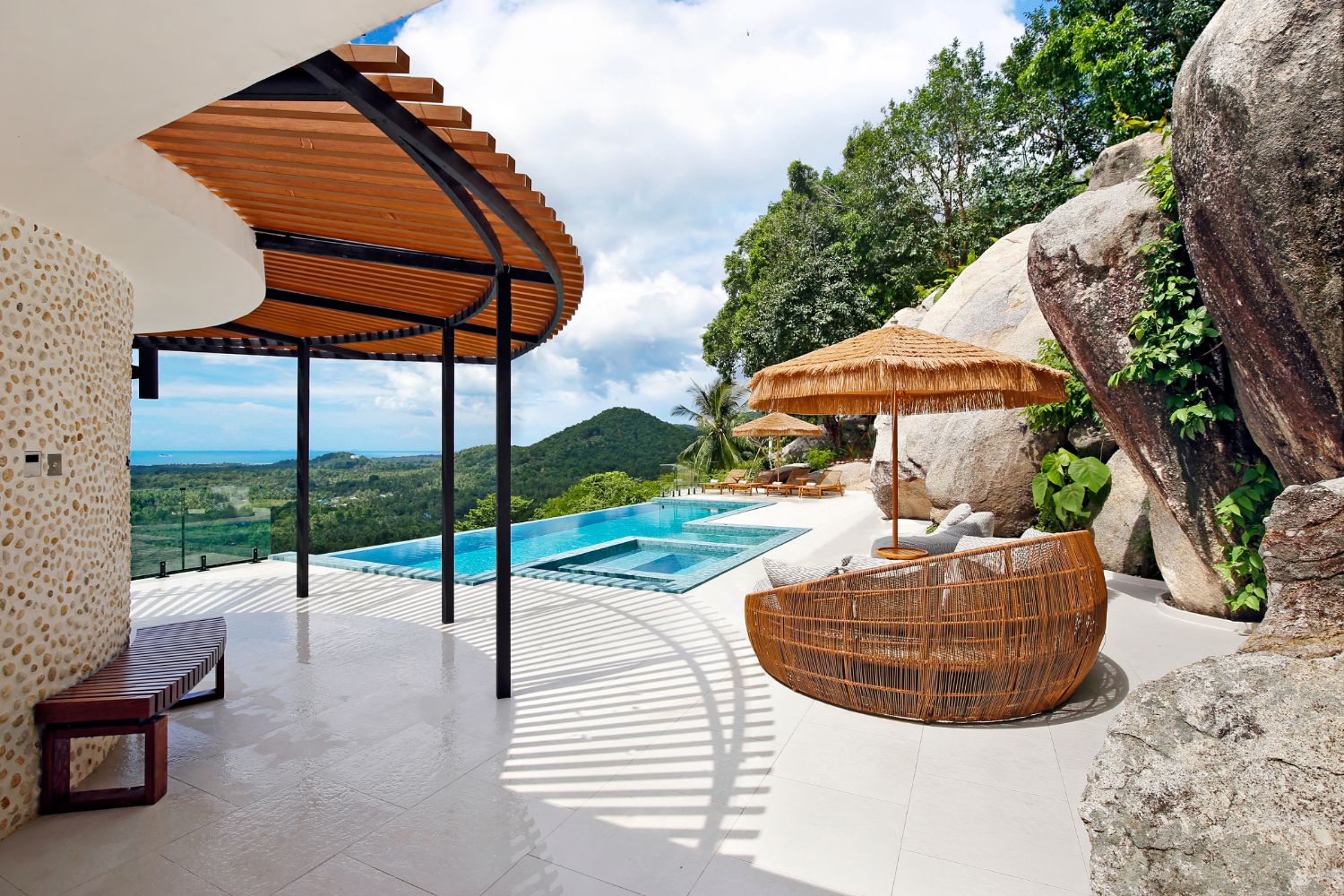 Poolside terrace with a thatched parasol and a round rattan chair between granite boulders, the sea and hills behind